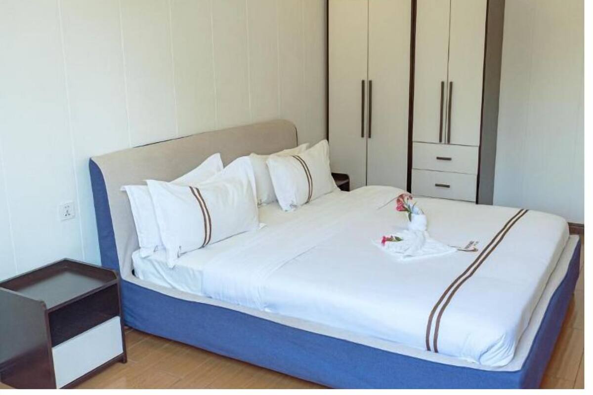 Una cama bien tendida con ropa de cama blanca y franjas marrones, una mesita de noche de madera y un armario con dos puertas.