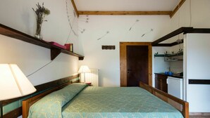 Una camera da letto con un letto, lampade da comodino, una testiera in legno, un comodino, uno scaffale a parete con libri e un vaso, una porta e un angolo cottura.