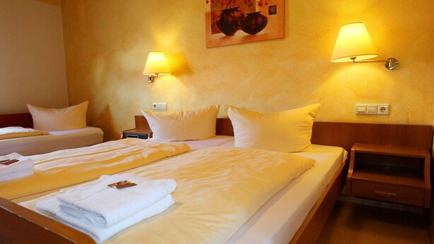 Habitación de hotel con dos camas, cada una con sábanas y almohadas blancas, una mesita de noche de madera, una lámpara de pared y un cuadro sobre las camas.