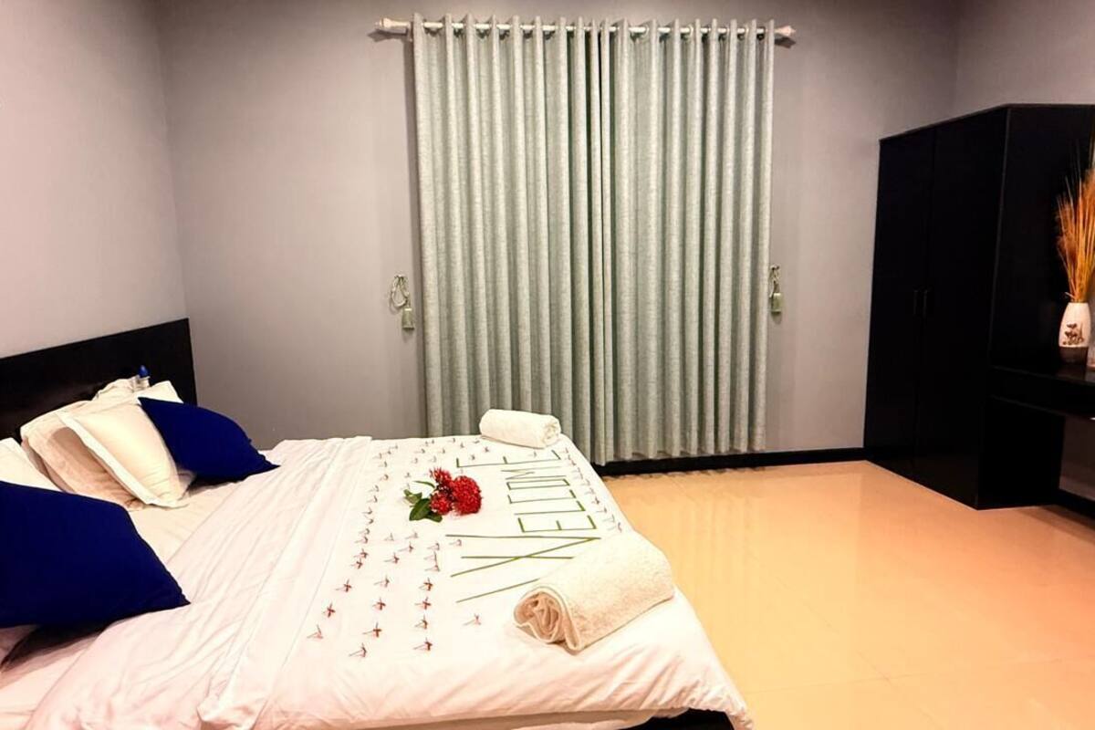 Ein ordentlich bezogenes Bett, eine Fußmatte, ein schwarzer Kleiderschrank und ein kleiner Tisch mit einer Vase.