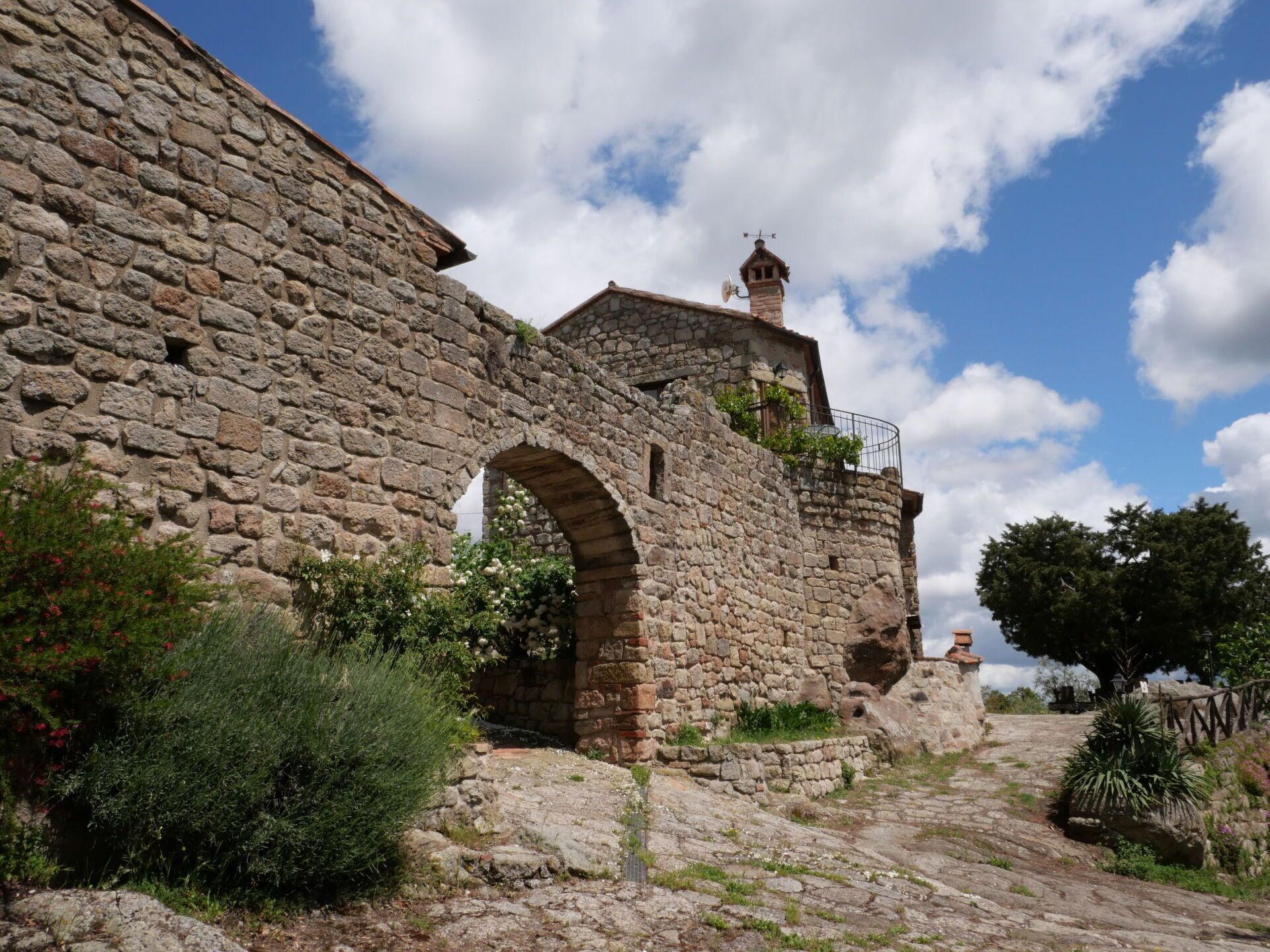 A stone building with an arched doorway, a small tower with a weather vane, and a clear blue sky with clouds.