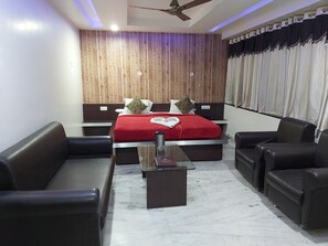 Una habitación de hotel moderna con una pared decorada con madera, una cama grande con ropa de cama roja, dos sofás de cuero negro, una mesa de centro de vidrio y un ventilador de techo.