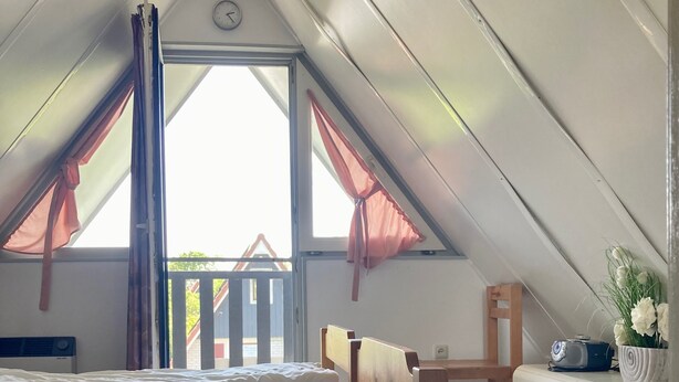 Ein Zimmer mit schrägen Wänden, einem Bett mit weißer Bettwäsche, einem Holzstuhl, einer weißen Kommode und einem Fenster mit Vorhängen.