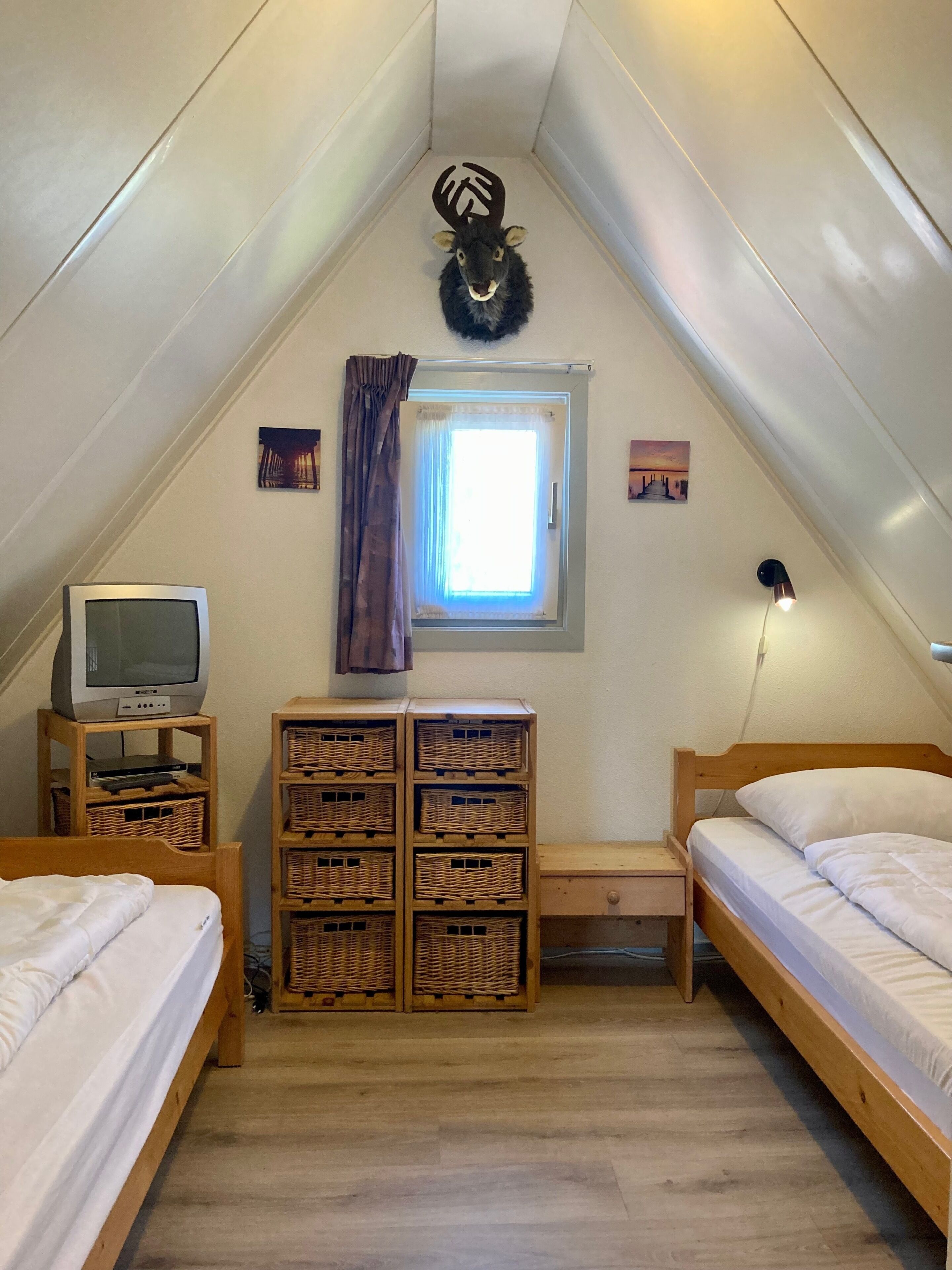 A small, cozy attic bedroom with two wooden beds, a television on a stand, wicker baskets, and a window with curtains.