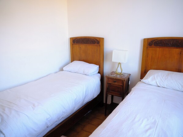 Two single beds with wooden headboards, a bedside table with a lamp, and a plain white wall.