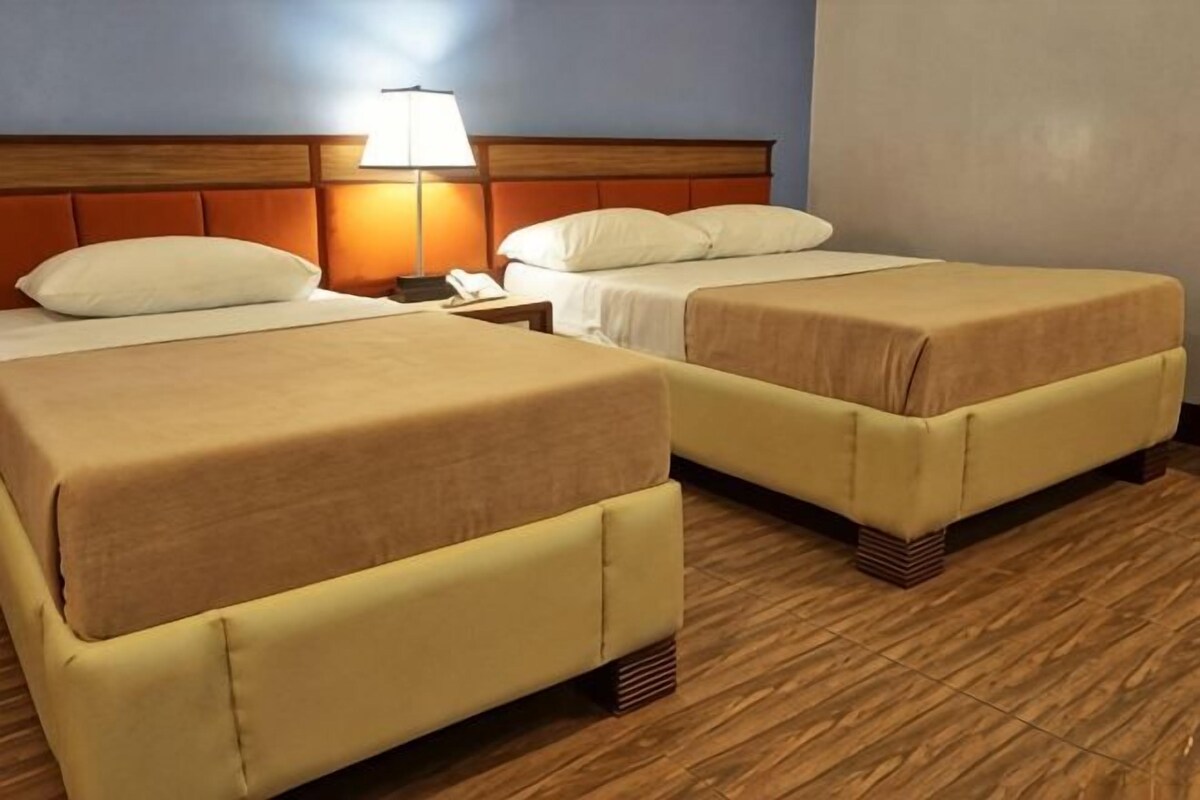 Una habitación de hotel con dos camas, una mesita de noche, una lámpara y suelo de madera.