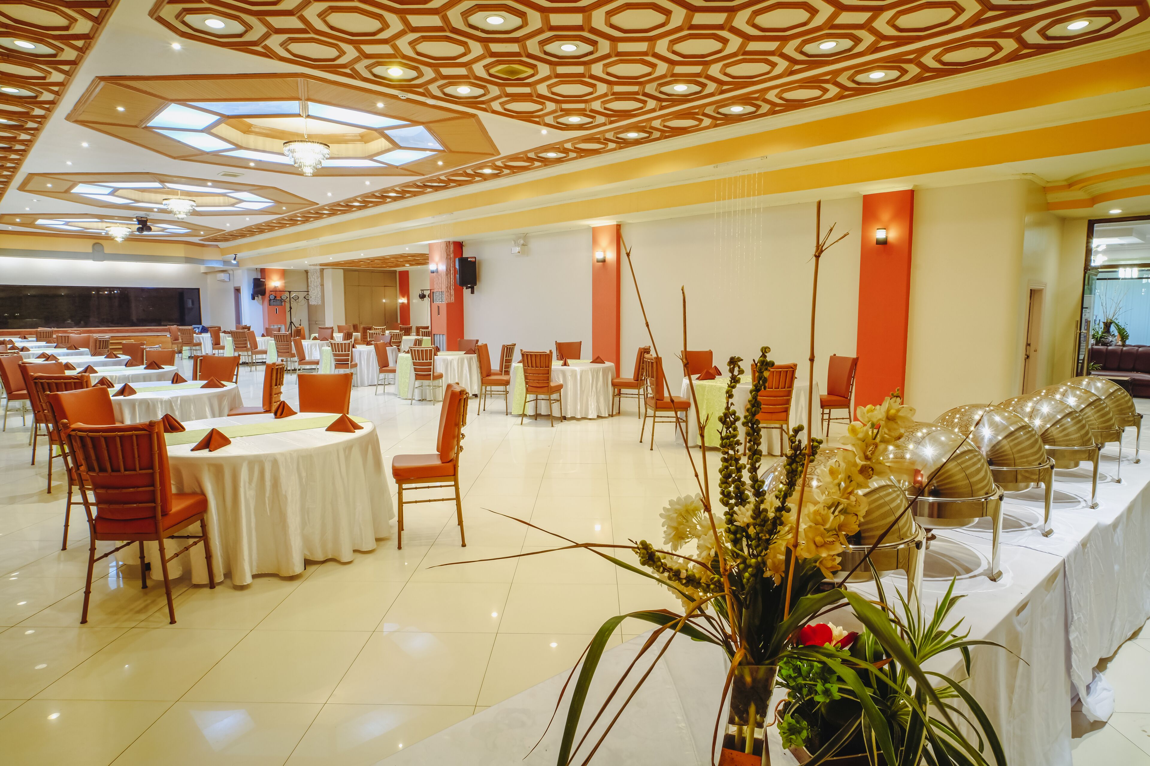 A spacious banquet hall with round tables, orange chairs, and a decorative ceiling.