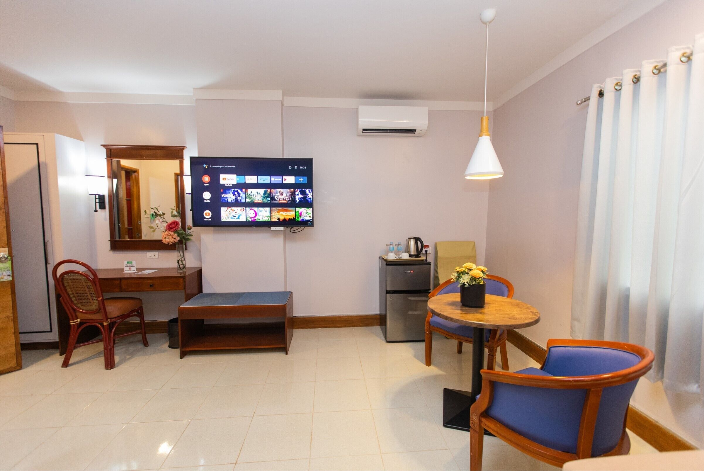 A hotel room with a flat-screen TV mounted on the wall, a wooden desk, a dining table with two chairs, a small refrigerator, and a ceiling-mounted air conditioner.