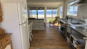 Una cocina con suelo de madera, armarios blancos, un refrigerador, un microondas y vistas a la playa a través de amplios ventanales.