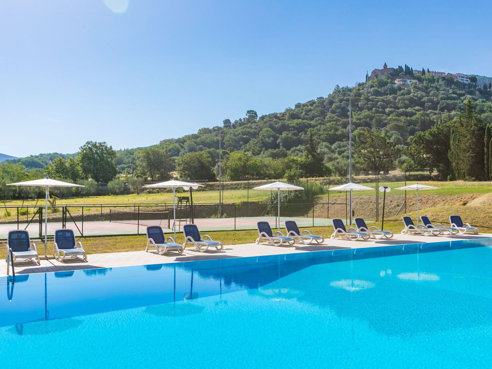 A swimming pool with lounge chairs and umbrellas, surrounded by a fence, with a view of a hill and trees in the background.