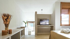 Une chambre d’hôtel moderne dotée d’une télévision à écran plat, d’un lit recouvert d’une housse beige, d’un plancher en bois et d’un vase contenant des fleurs séchées, posé sur une console blanche.