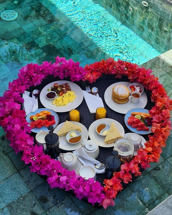 A heart-shaped table with various dishes and drinks, surrounded by vibrant pink flowers, set on a poolside area. - Ama-Lurra Villa (Gili Air)