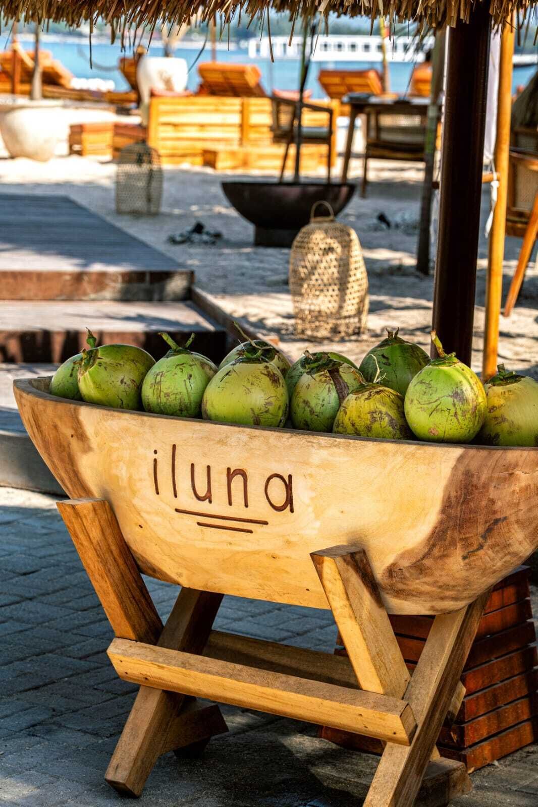 A wooden trough labeled 'iluna' filled with coconuts, placed on a paved area with lounge chairs and a thatched roof structure in the background.