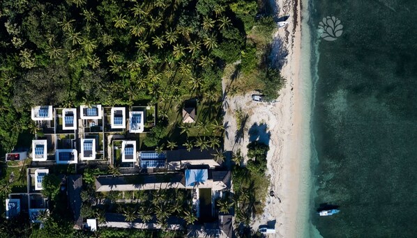 An aerial view of a resort with multiple square buildings, some with solar panels, adjacent to a sandy beach and surrounded by dense palm trees. - Ama-Lurra Villa (Gili Air)