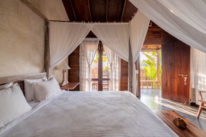 Une chambre avec un grand lit, une tête de lit en bois et une vue sur une terrasse en bois et des palmiers à l’extérieur.