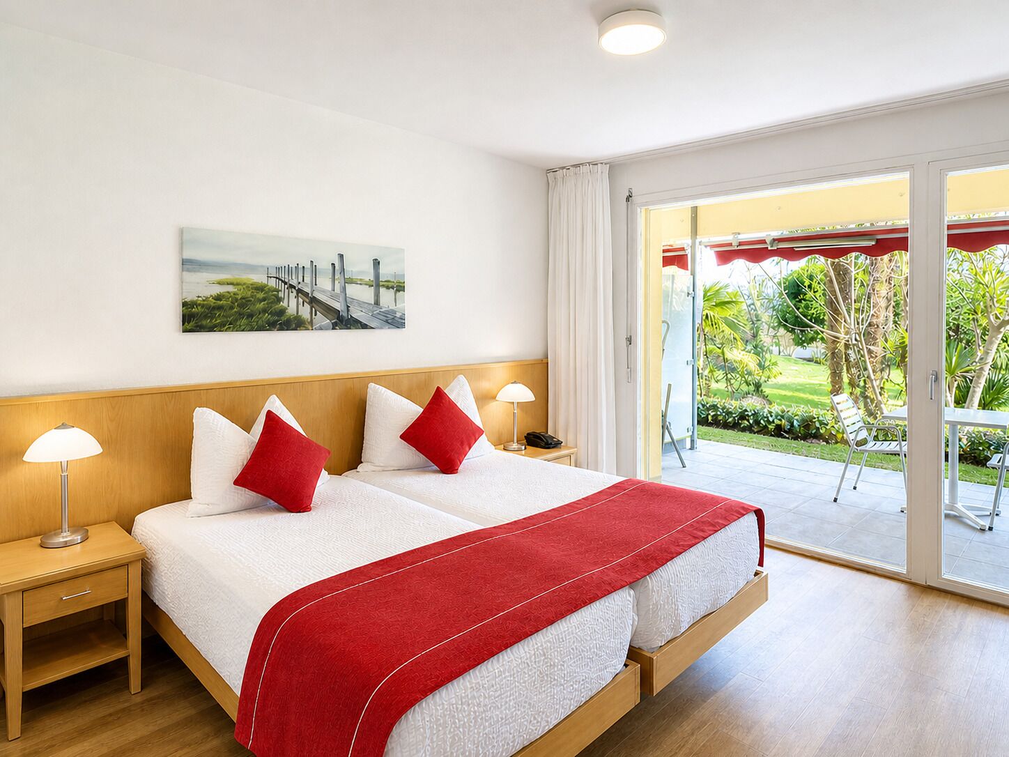 A double bed with white and red bedding, two bedside tables with lamps, a painting on the wall, and a view of a garden through a sliding door.