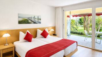 A double bed with white and red bedding, two bedside tables with lamps, a painting on the wall, and a view of a garden through a sliding door.
