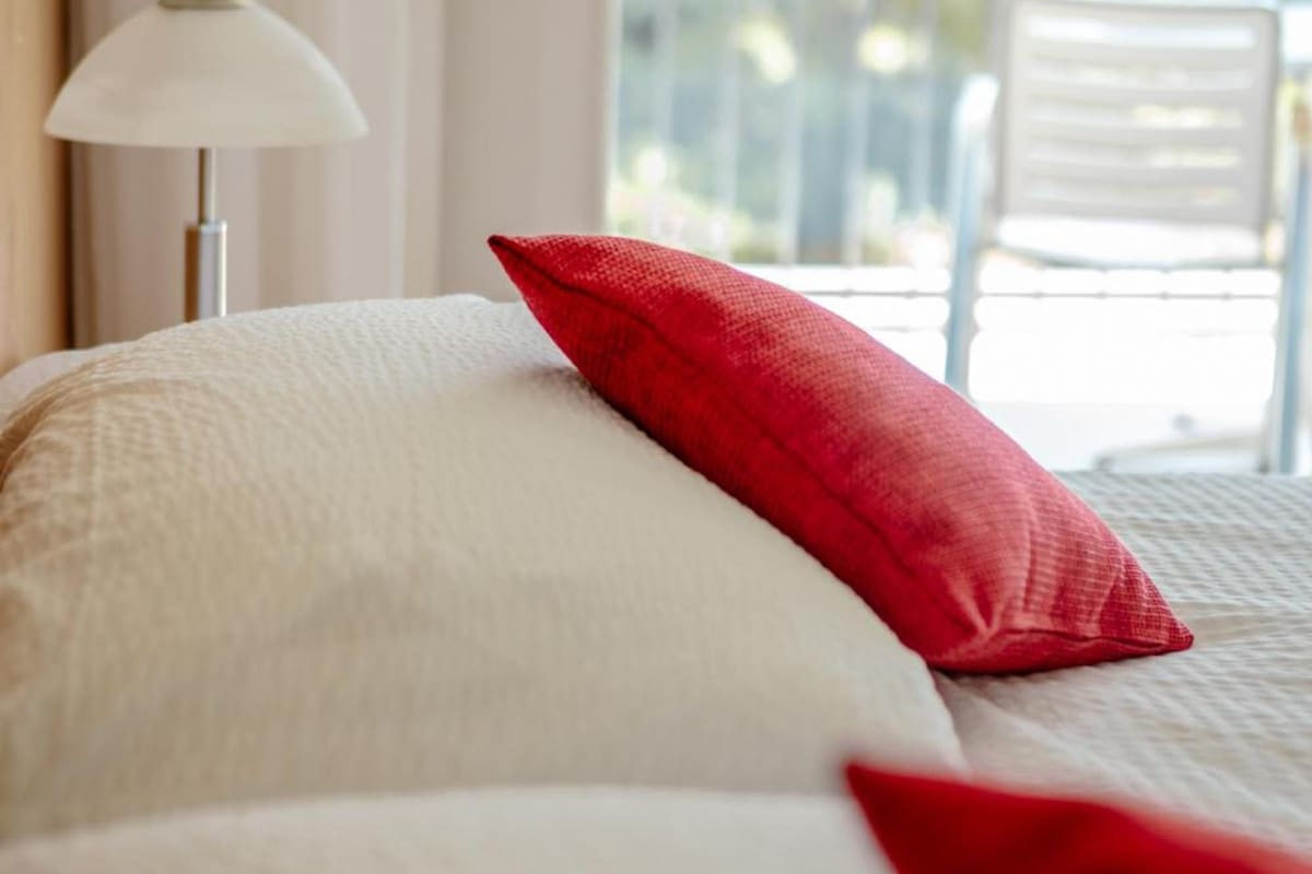 A bed with white bedding and a red pillow, a white lamp, and a view of a balcony with a chair.