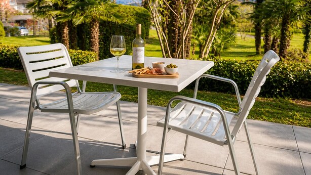 Une table avec une bouteille de vin, un verre et une assiette de nourriture, entourée de chaises, est placée sur une terrasse d’où l’on peut admirer un paysage verdoyant et des montagnes.