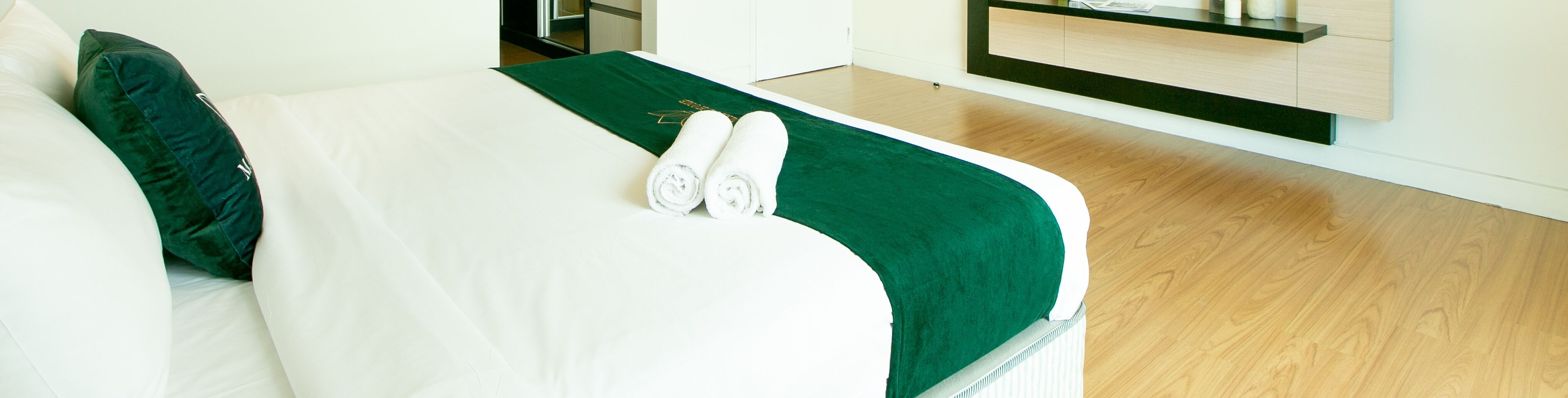 A neatly made bed with white linens and a green accent, a wooden floor, and a minimalist room design.
