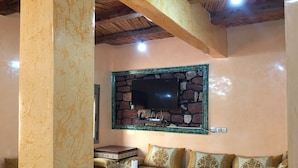 Una sala acogedora con un televisor instalado en una pared de piedra, una mesa redonda con mantel blanco y un sofá con cojines estampados.