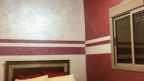Un dormitorio con techo de madera, cama con cabecera estampada y ventana con celosía decorativa.