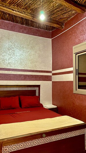 Ein Zimmer mit einem Bett, einem Nachttisch, einem Fenster und einer Holzbalkendecke.
