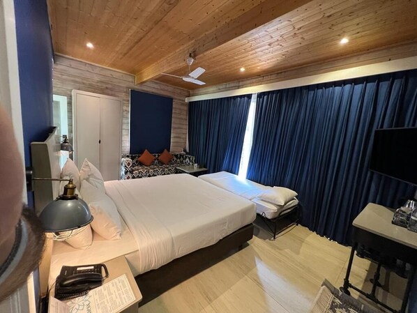A double bed with white linens, a desk with a lamp, a telephone, and a small table with a bottle.