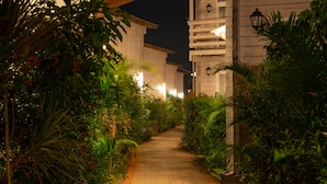 A pathway flanked by lush greenery and wooden buildings with exterior lighting.