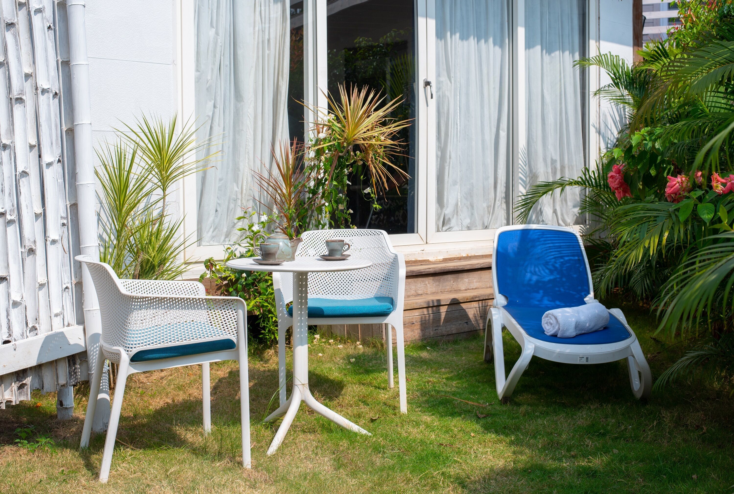 Um pátio com duas cadeiras brancas, uma delas com almofada azul, e uma pequena mesa redonda com tampo branco.