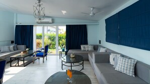A spacious living room with a large sofa set, a coffee table, and a chandelier. There is a sliding glass door leading to a balcony with a view of greenery.