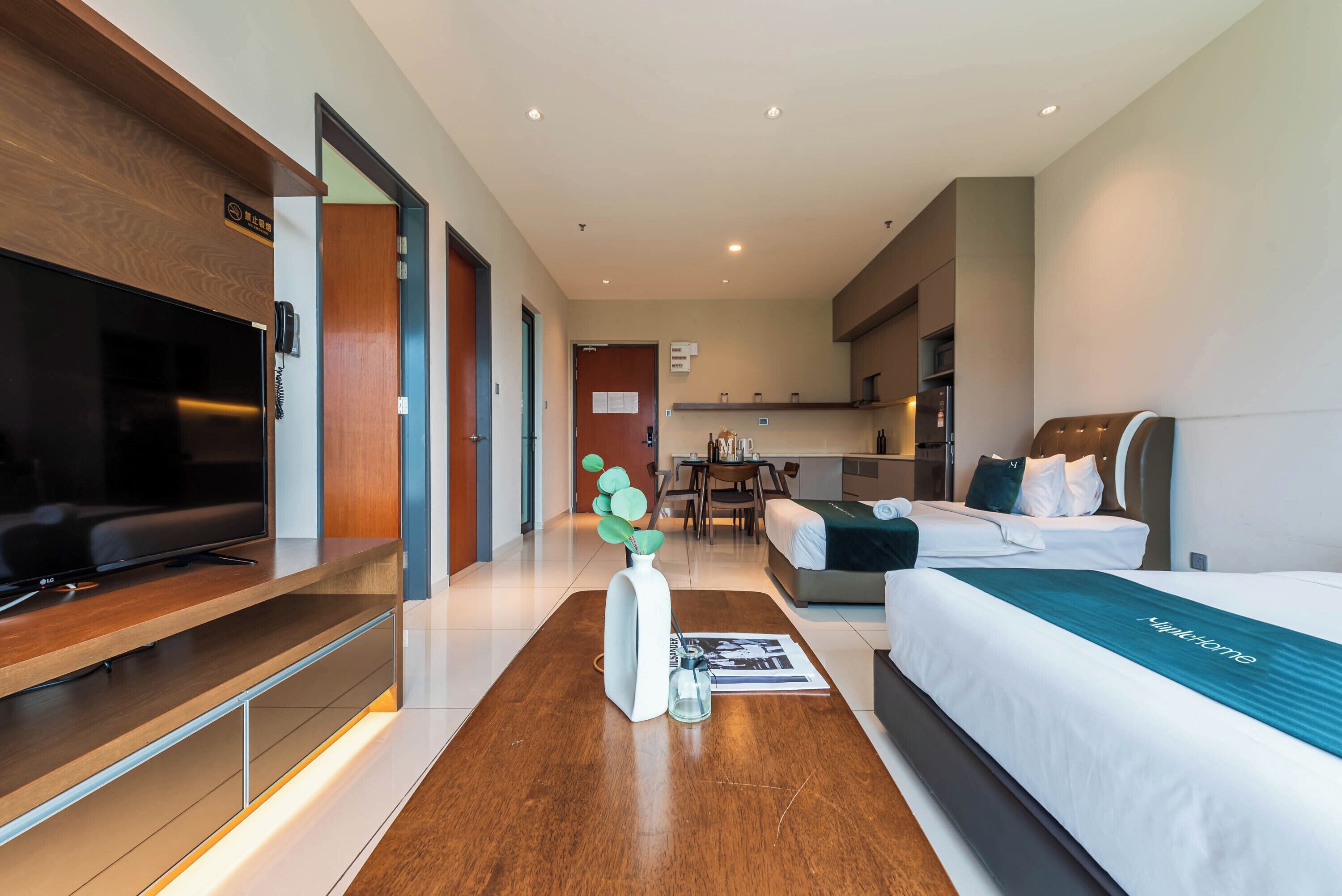 A hotel room with a flat-screen TV, a bed with a teal bedspread, a sofa, a dining table with chairs, and a small kitchen area.