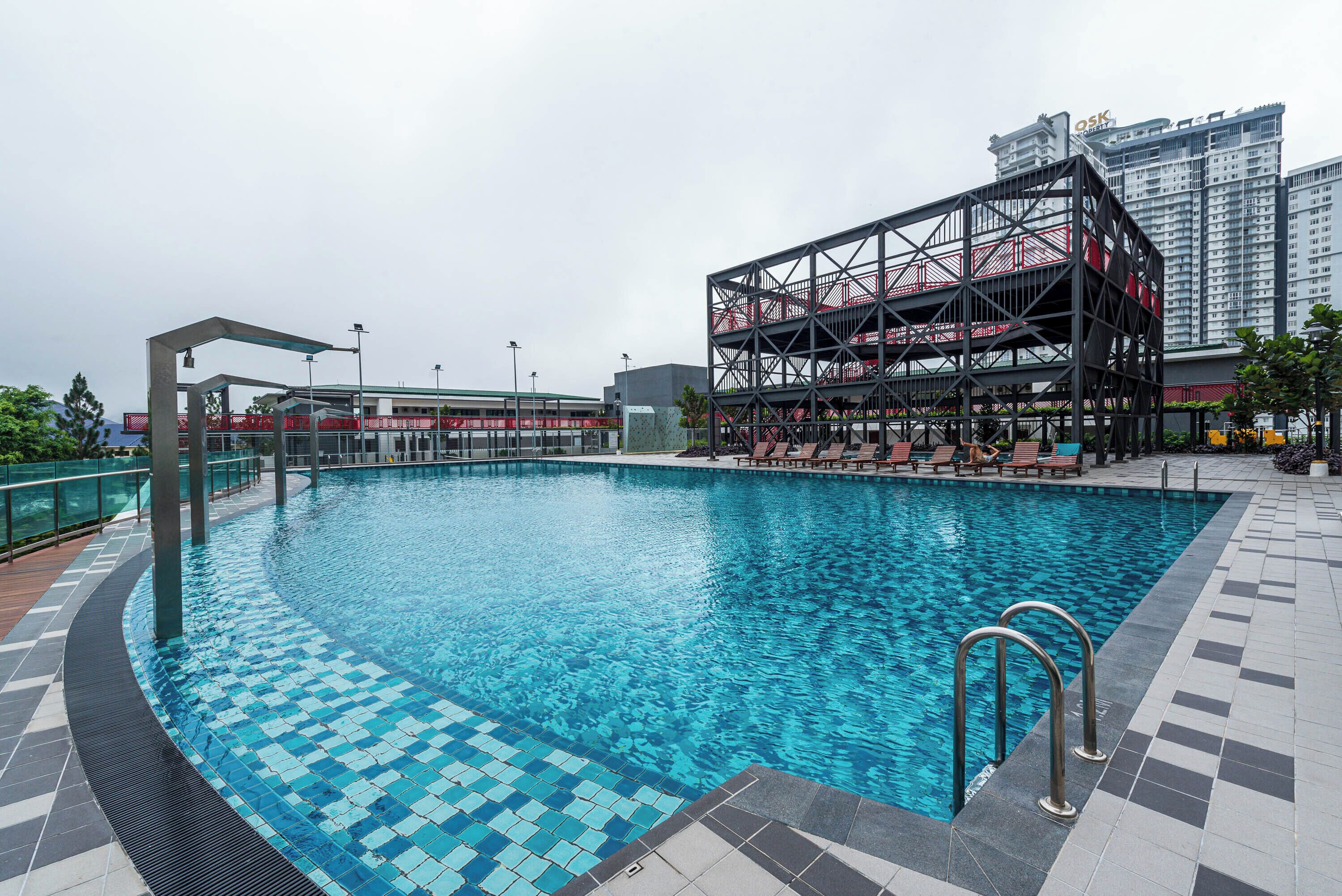 Uma piscina com um deck ladrilhado, rodeada por uma estrutura metálica com guarda-corpos vermelhos, e um prédio alto ao fundo.