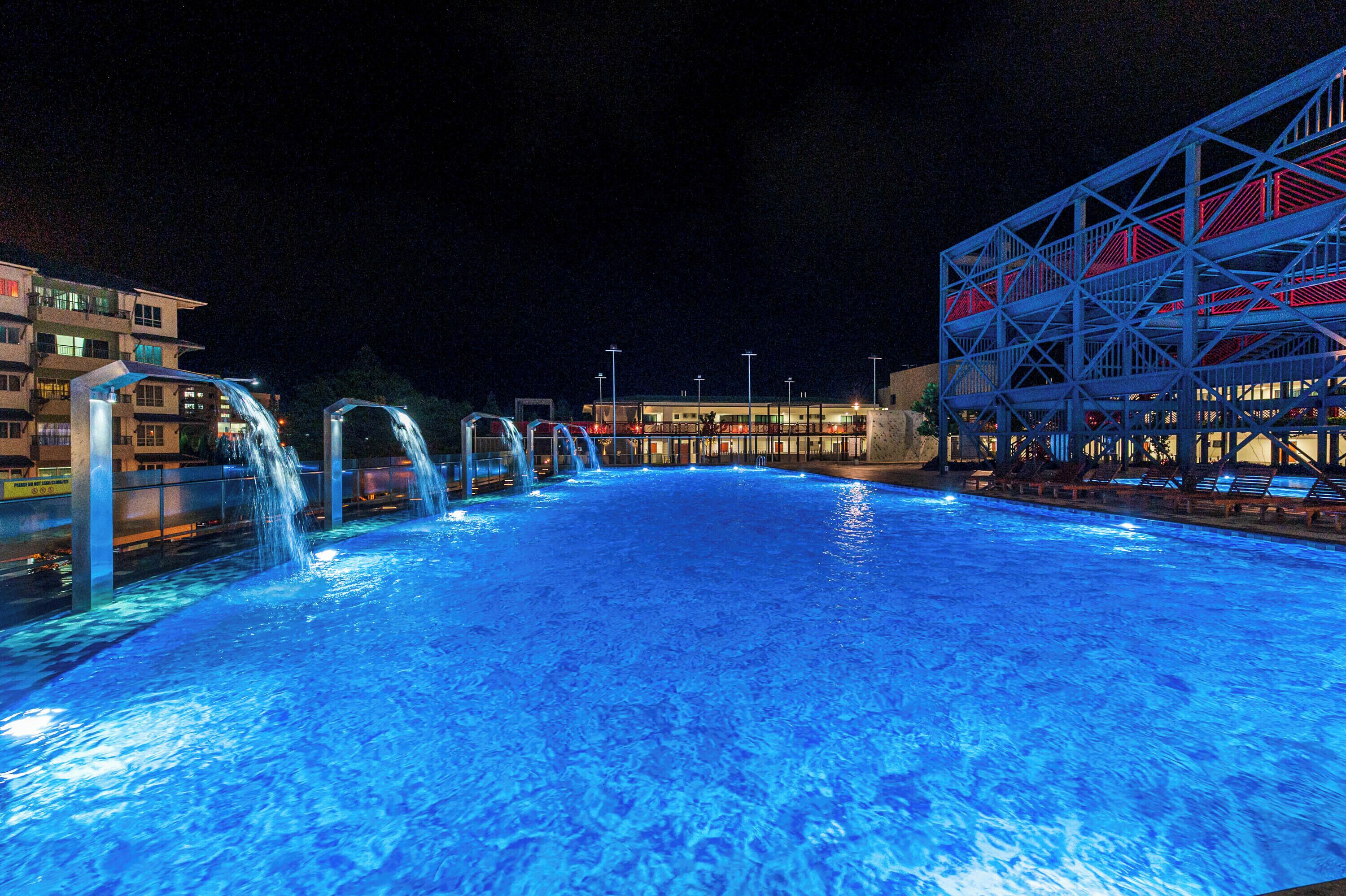 Une piscine nocturne agrémentée de jeux d’eau, avec un bâtiment illuminé en rouge et en bleu en arrière-plan.