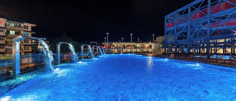 A nighttime swimming pool with water features and a building with red and blue lighting in the background.
