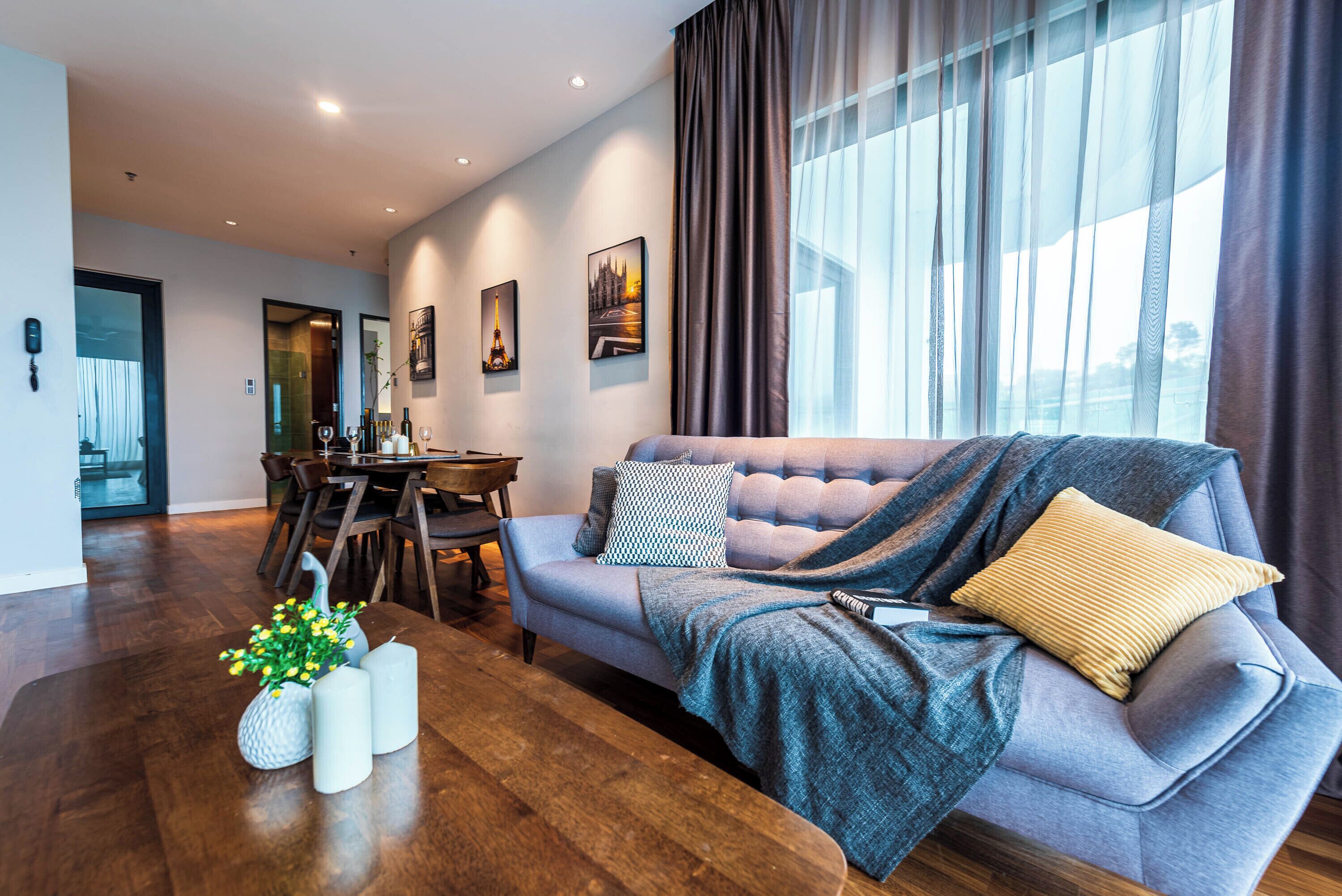 A modern living room with a grey sofa, a dining table, and a large window with curtains.