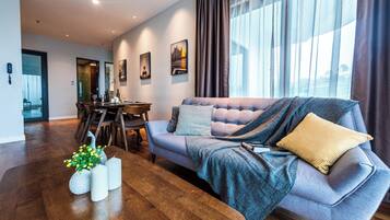 A modern living room with a grey sofa, a dining table, and a large window with curtains.