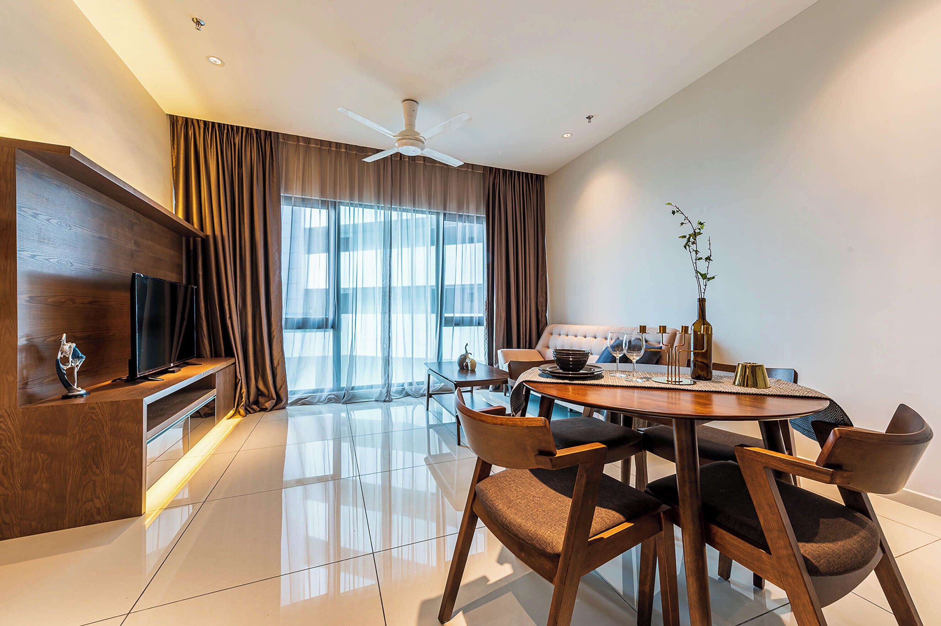 A modern living room with a flat-screen TV, a dining area with a round table and chairs, and a large window with curtains.