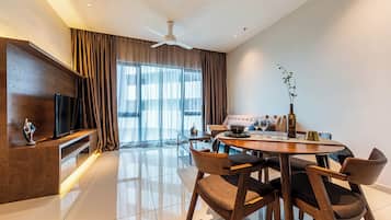 A modern living room with a flat-screen TV, a dining area with a round table and chairs, and a large window with curtains.