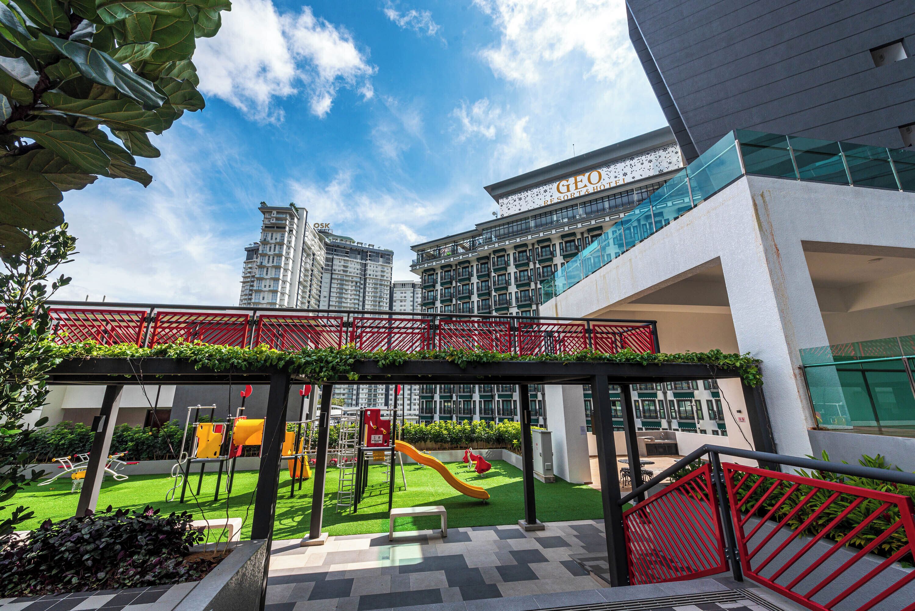 A playground with slides and climbing structures, surrounded by greenery and a red fence.