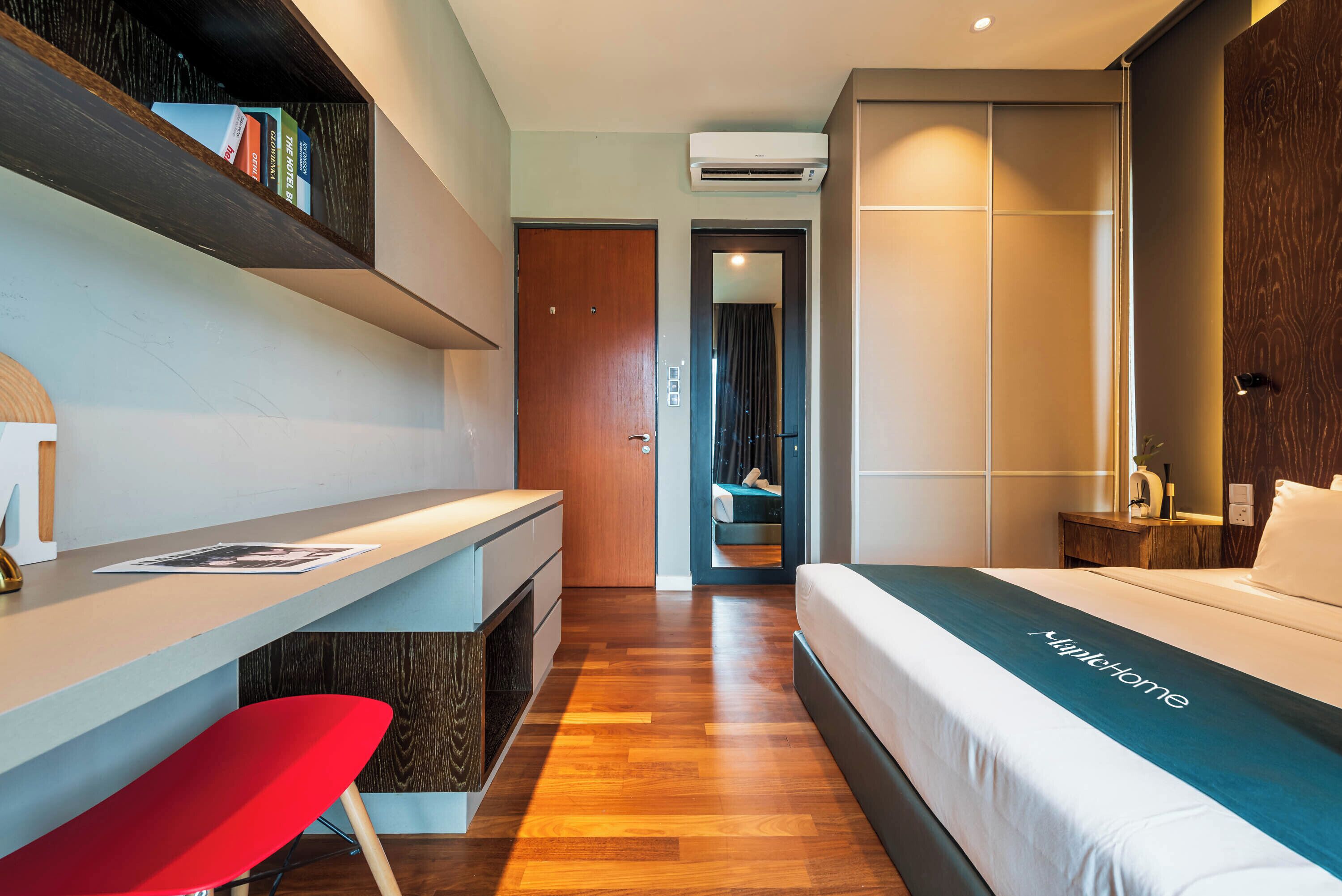 Quarto de hotel moderno com piso de madeira, cama com colcha verde-água, escrivaninha com cadeira vermelha e estante com livros.