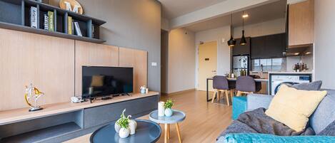 A modern living room with a TV, sofa, coffee table, and a dining area with chairs and a kitchen in the background.