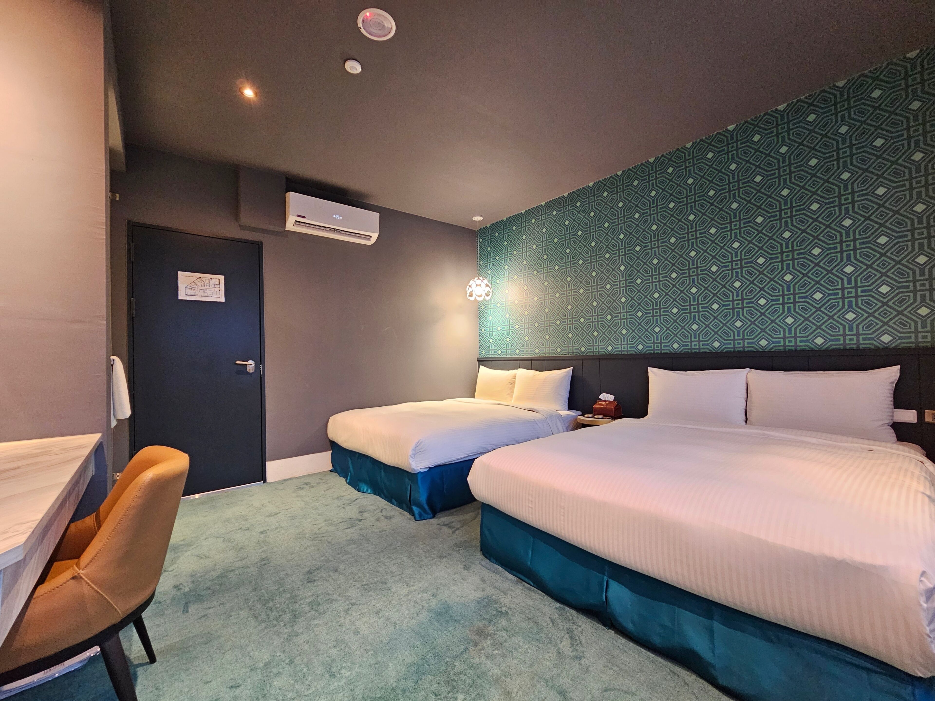 Habitación de hotel con dos camas, un escritorio, una silla, un equipo de aire acondicionado y un papel tapiz estampado.