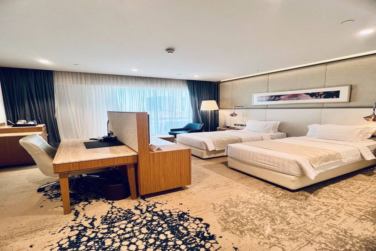 A hotel room with a desk, two beds, a chair, a lamp, and a large window with curtains.