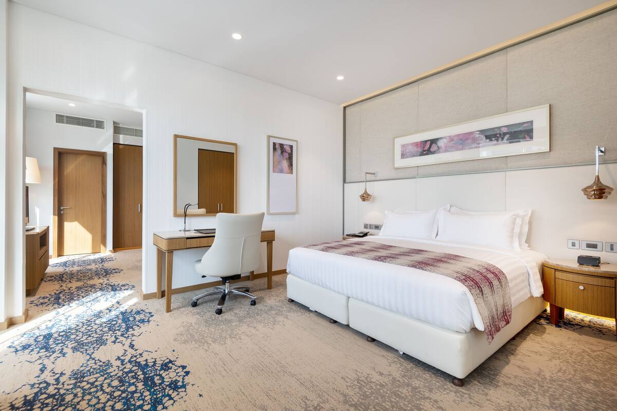 A hotel room with a large bed, a desk, a chair, a bedside table, a lamp, and a framed picture on the wall.