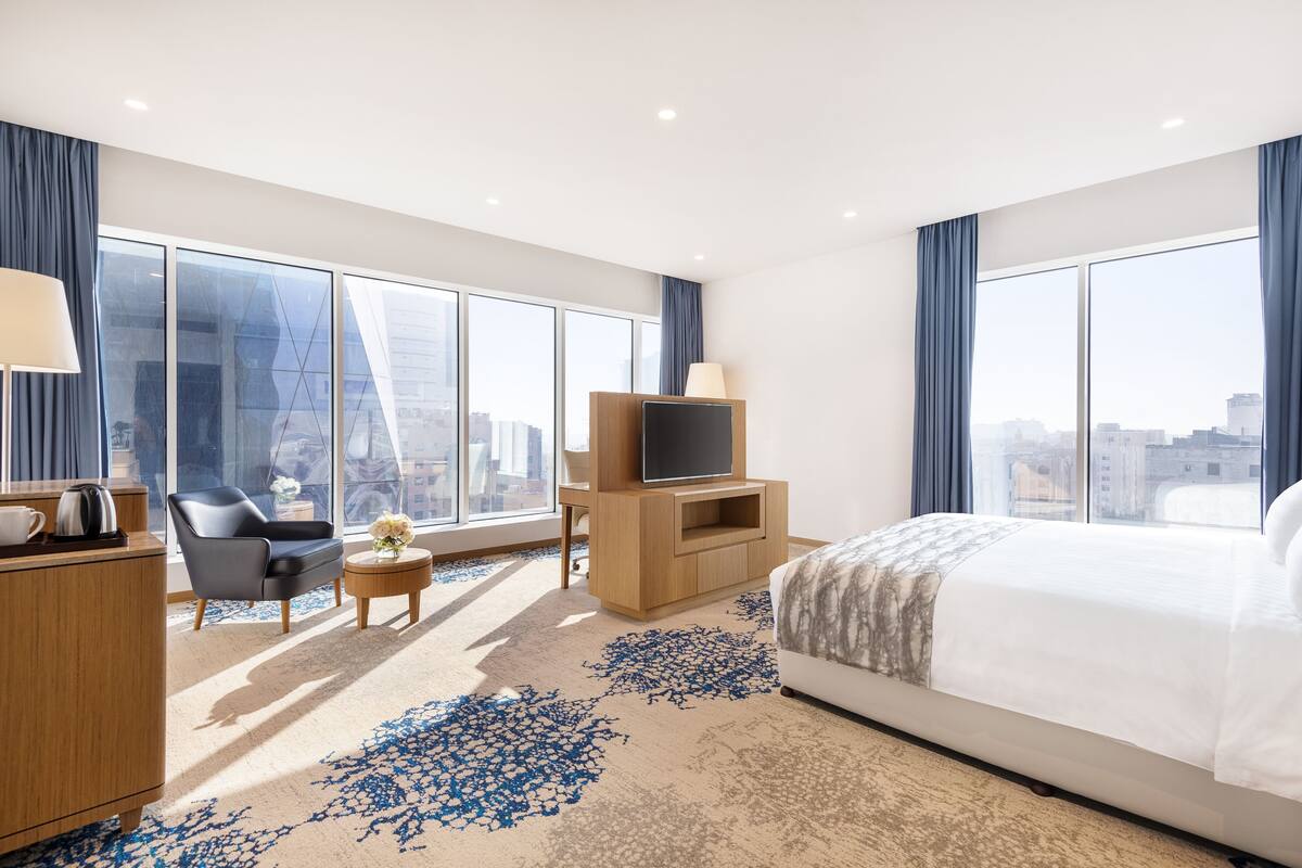 A modern hotel room with a large bed, a TV, a desk with a lamp, and a view of the city.