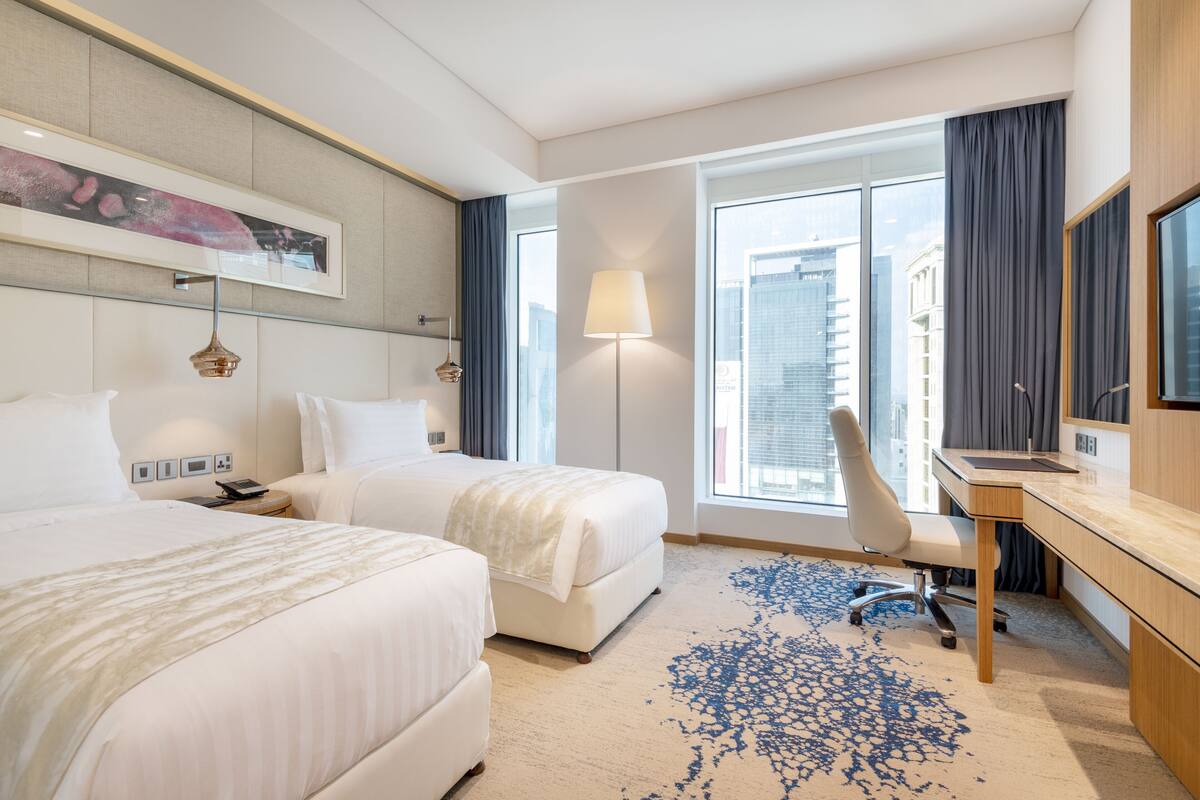 A hotel room with two beds, a desk, a chair, a lamp, and a large window with curtains.
