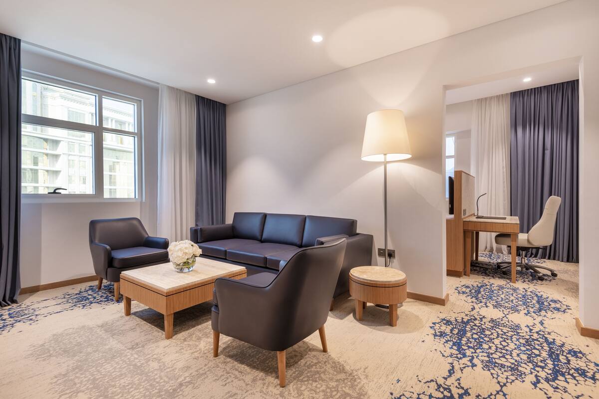 A modern hotel room with a sofa, armchairs, a coffee table, a desk, and a lamp.