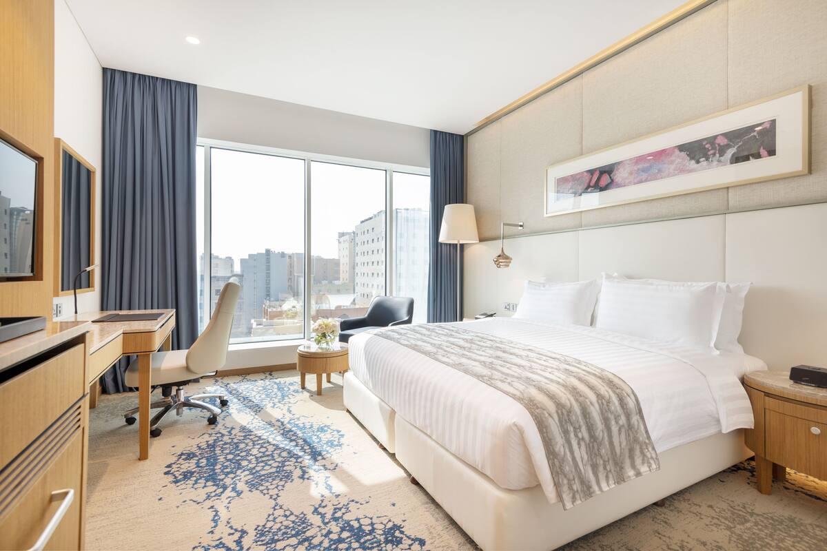 A hotel room with a large bed, a desk, a chair, a TV, and a window with a city view.
