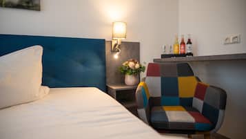 A hotel room with a bed, a colorful armchair, a wall-mounted lamp, a small table with a vase of flowers, and a shelf with bottles.